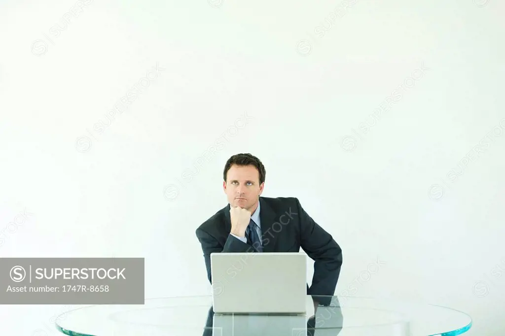 Businessman sitting at desk with laptop computer, hand under chin, looking up