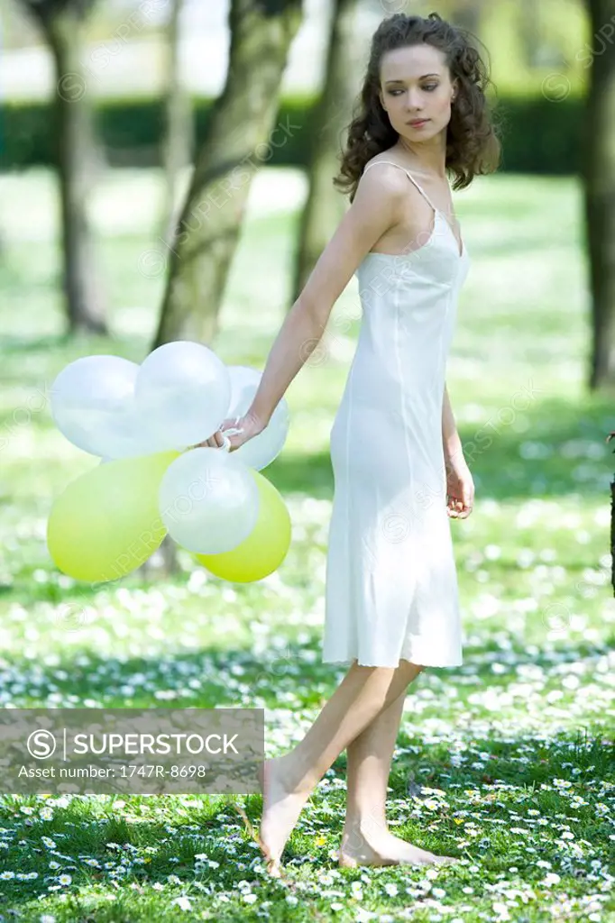 Young woman walking in field of flowers, holding balloons behind back, looking over shoulder