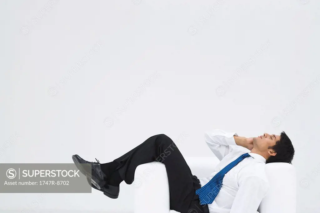 Businessman lying sideways in armchair, making phone call, full length