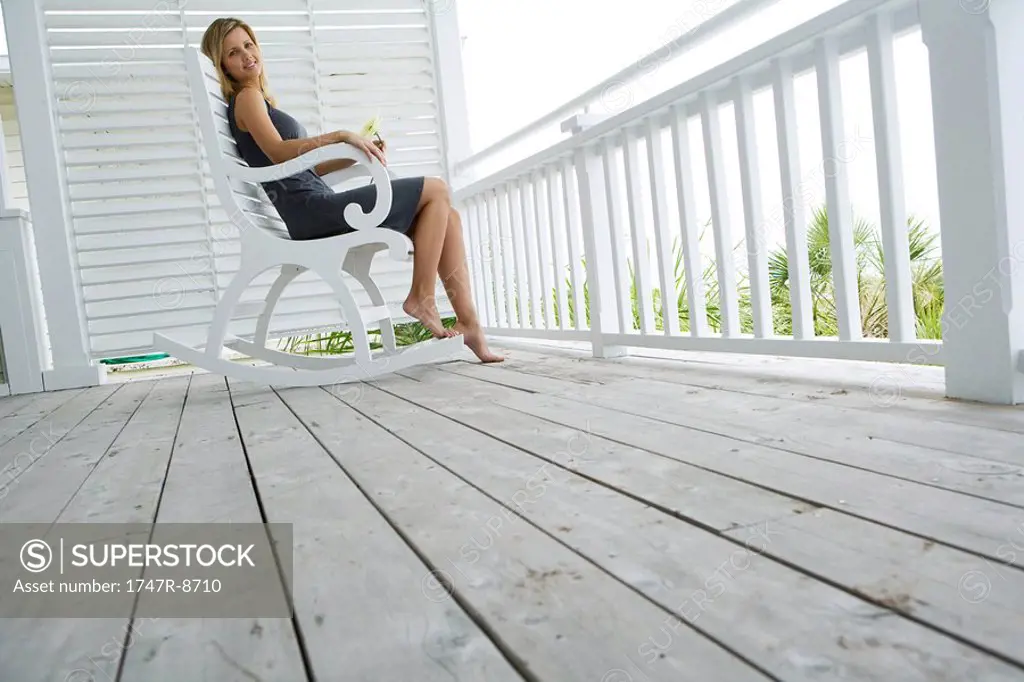 Woman sitting in rocking chair on porch, holding flower, smiling at camera