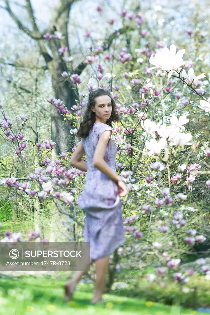 Young woman in dress walking beside flowering tree, looking over shoulder at camera