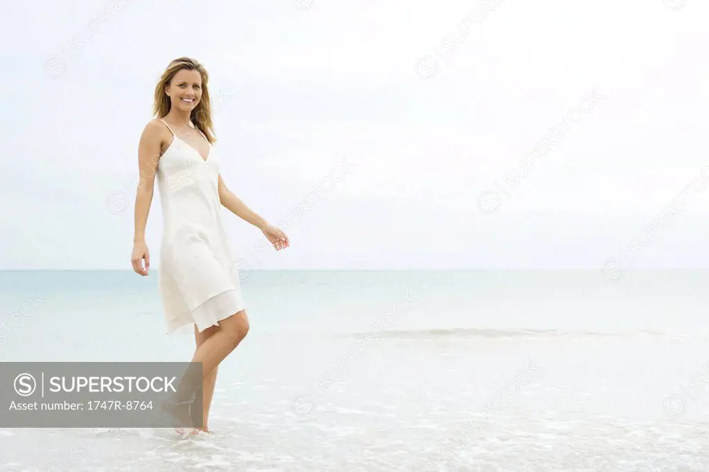 Young woman in sundress walking in shallow water, smiling at camera