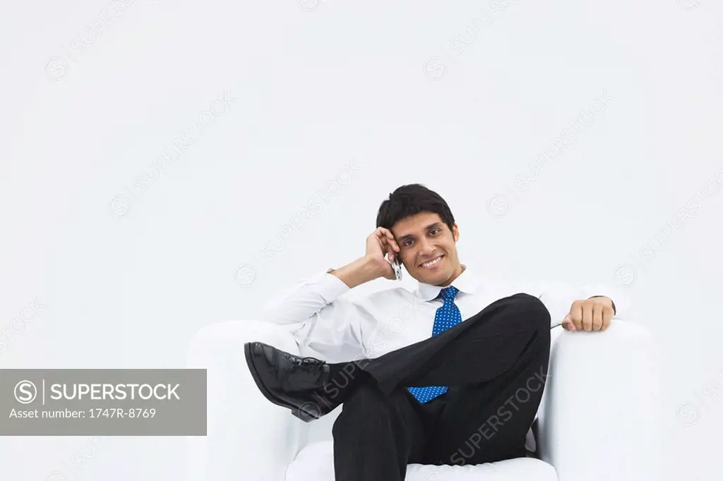 Businessman sitting in armchair, using cell phone, smiling at camera