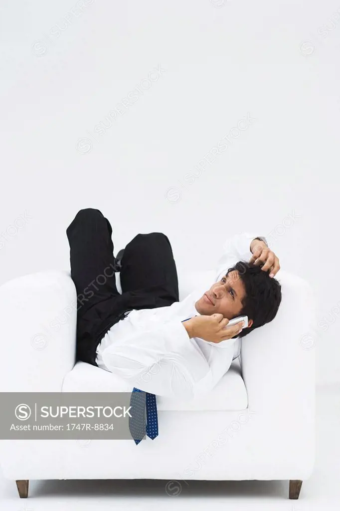 Businessman lying backwards in armchair, holding cell phone to ear and scratching head, full length