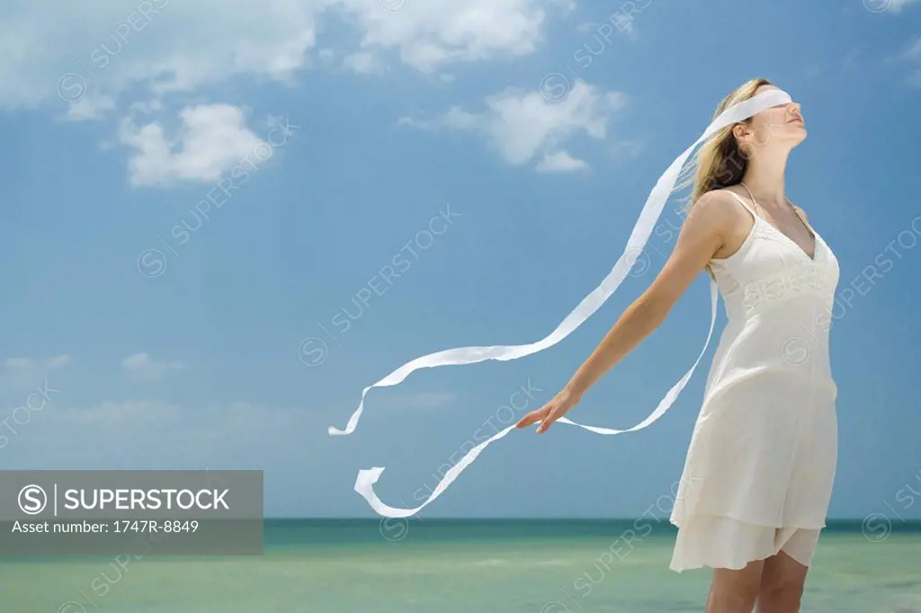 Young woman standing by the sea with ribbon covering eyes, tousled by breeze, side view
