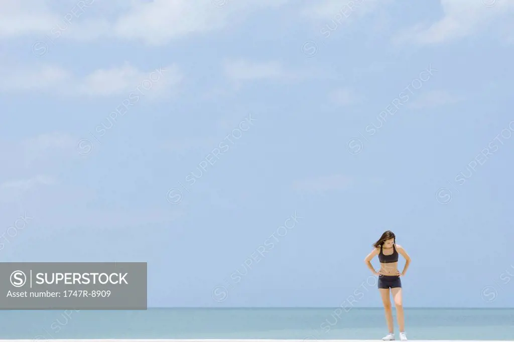 Young woman in sports clothing standing at the beach, looking down, mid-distance