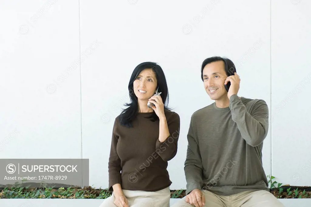 Couple sitting side by side, both using cell phones, smiling