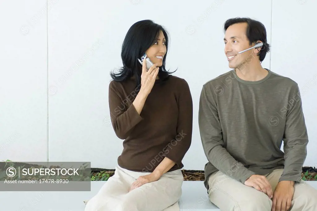 Couple sitting, smiling at each other, woman using cell phone, man wearing headset