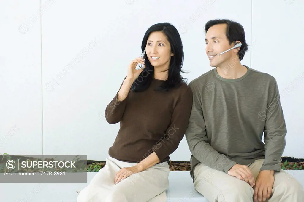 Couple sitting side by side, woman using cell phone, man wearing headset, both smiling