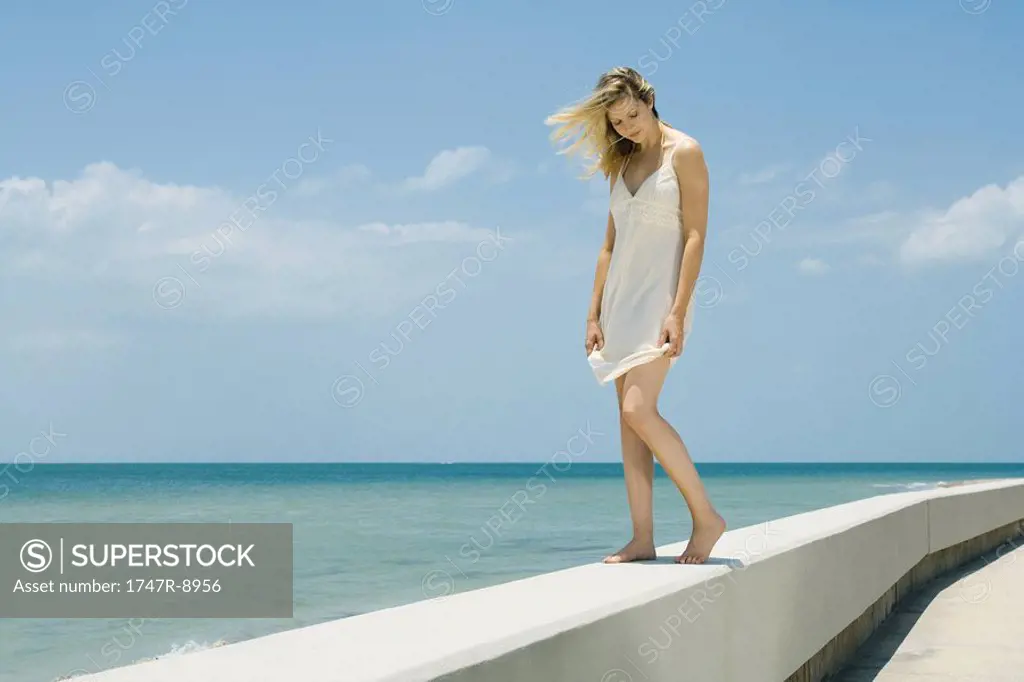 Young woman in sundress standing on low wall by the sea, eyes closed