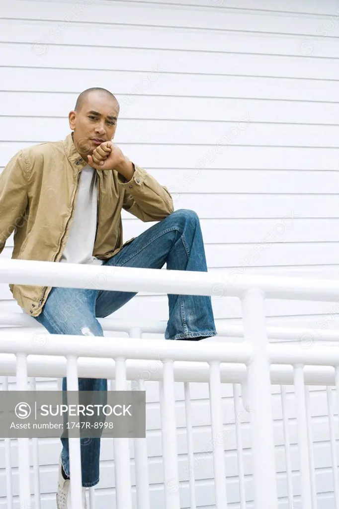 Casually dressed man sitting on railing, hand under chin, looking at camera