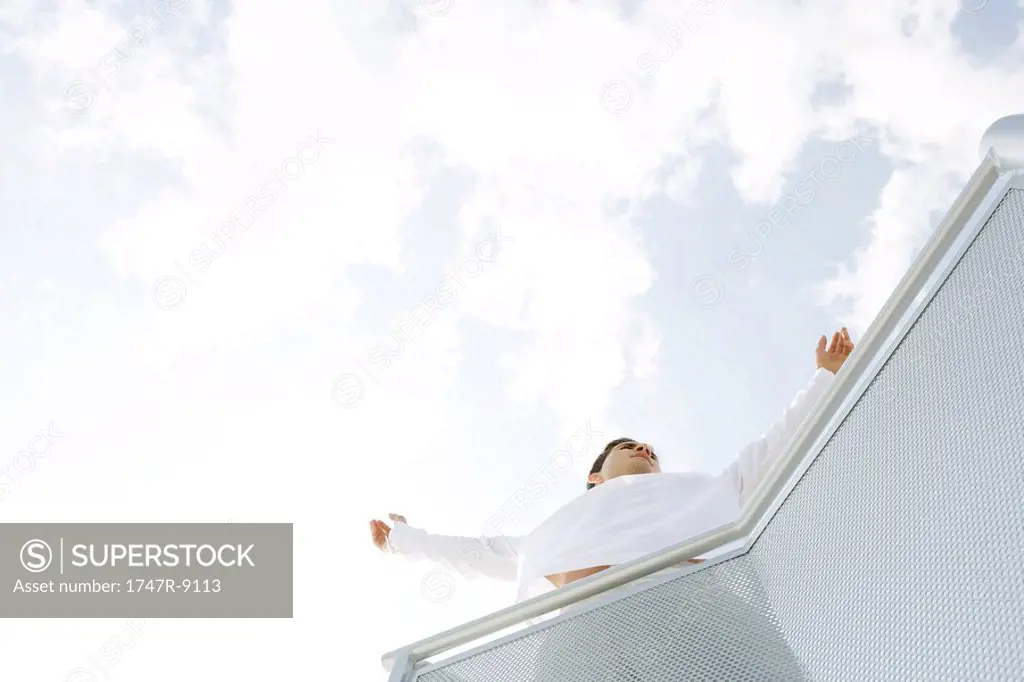 Man standing on balcony with arms out, sky in background, low angle view