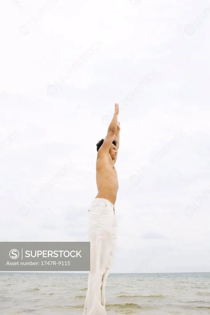 Man standing in front of ocean, stretching arms overhead, side view