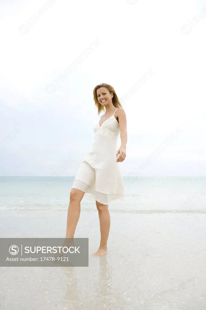 Young woman in sundress walking on wet sand at beach, smiling at camera, full length