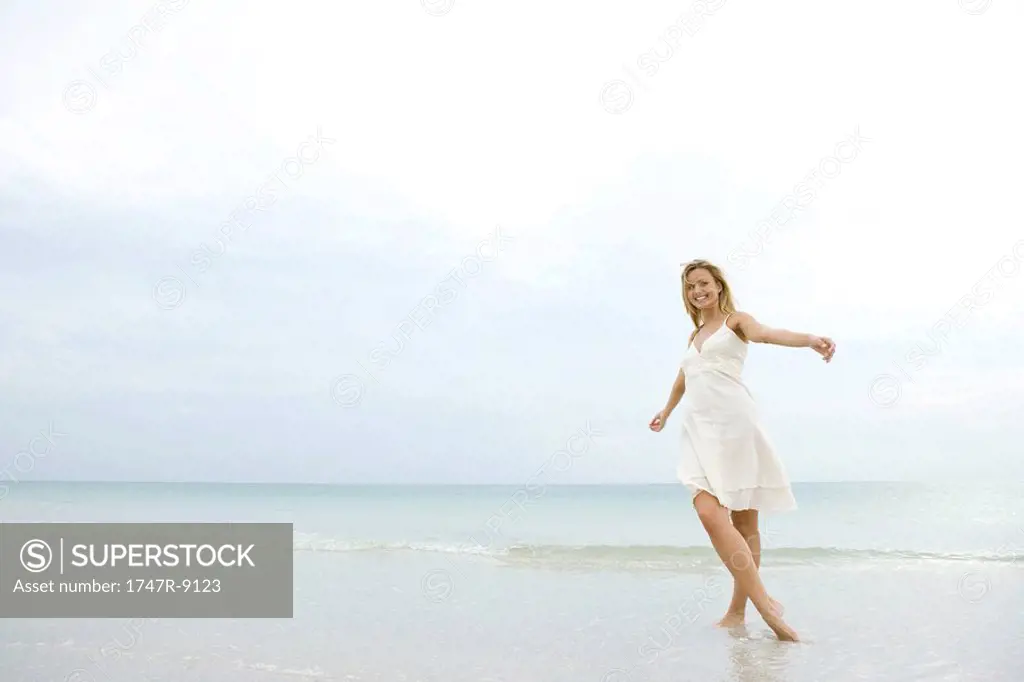 Young woman in sundress posing on beach with arms out and legs crossed, smiling at camera