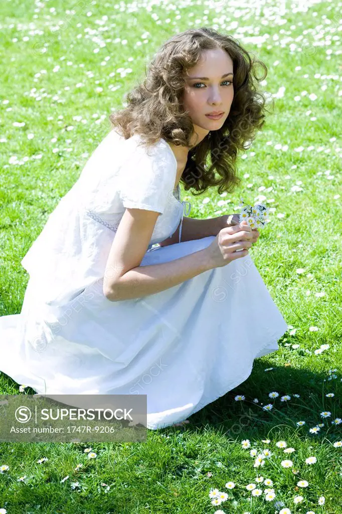 Young woman crouching in meadow, picking flowers, looking up at camera