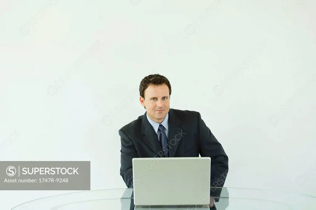 Businessman sitting at desk, using laptop computer, smiling at camera