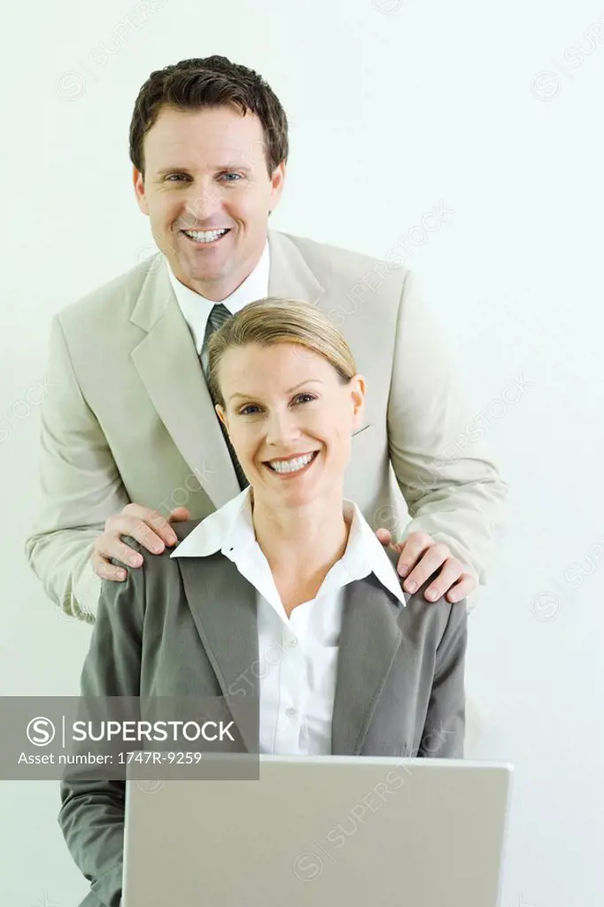 Businessman standing behind female associate with his hands on her shoulders, both smiling at camera