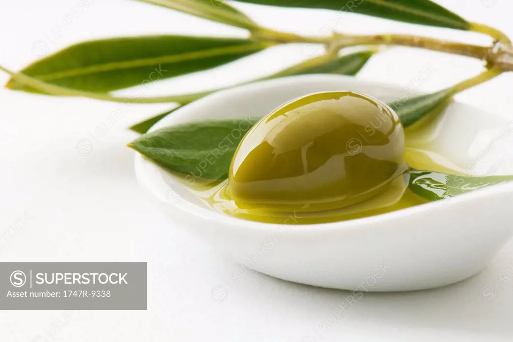 Green olive and sprig of leaves in small dish of olive oil, close-up