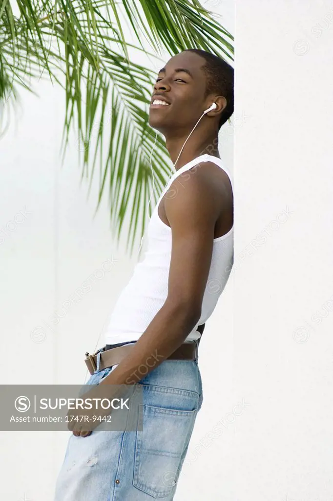Teen boy leaning against wall, listening to earphones, palm leaves in background
