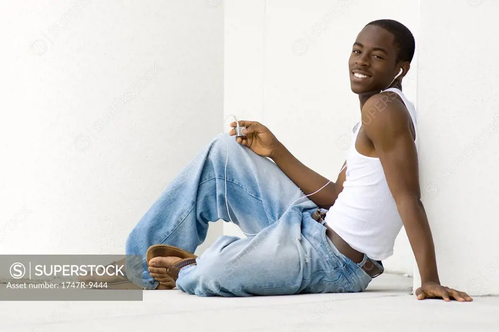 Teen boy sitting on floor, listening to MP3 player, smiling at camera, full length
