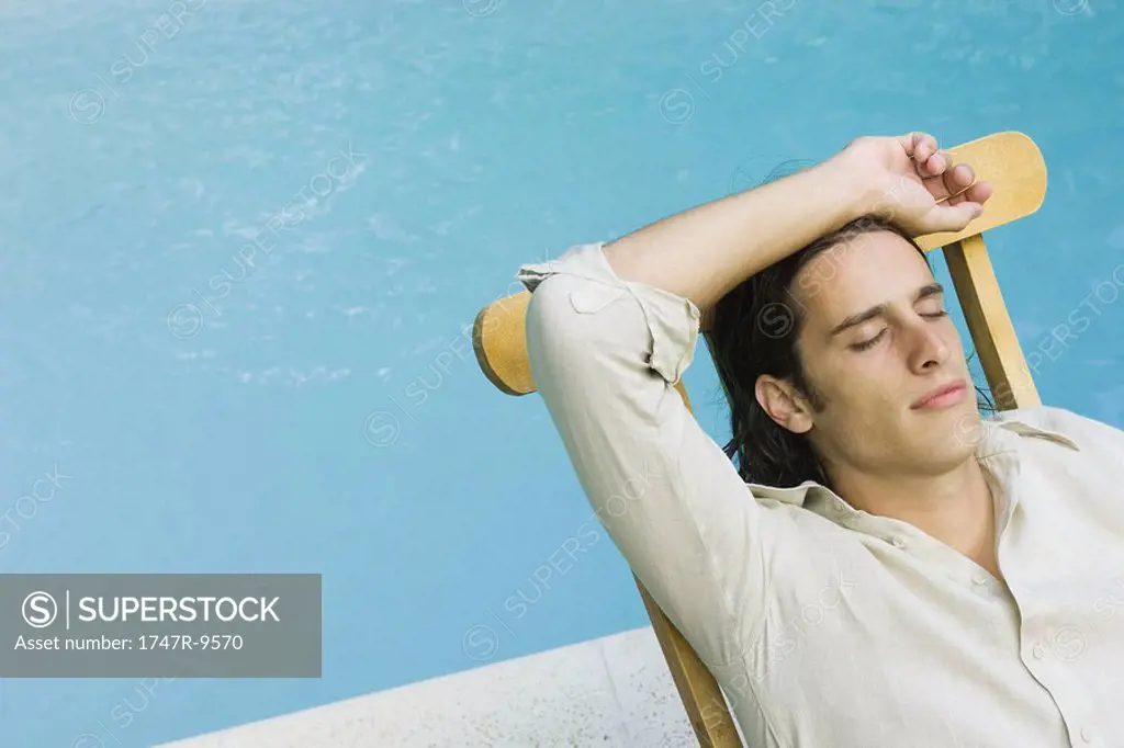 Man sitting in lounge chair beside swimming pool, eyes closed, high angle view