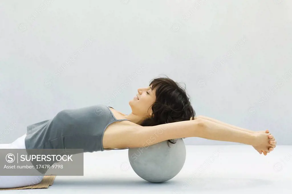 Woman leaning back against fitness ball, arms outstretched, side view