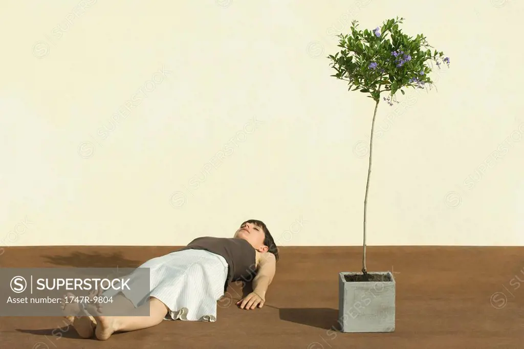 Woman lying on the ground beside potted tree, eyes closed