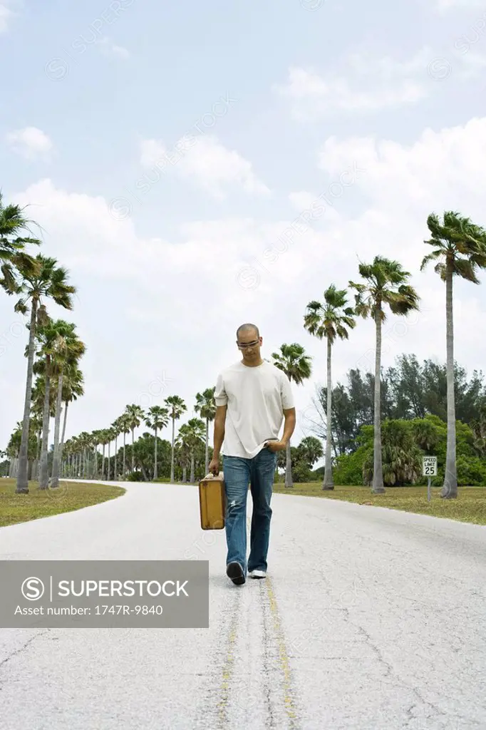 Man walking in center of road, carrying suitcase, looking down