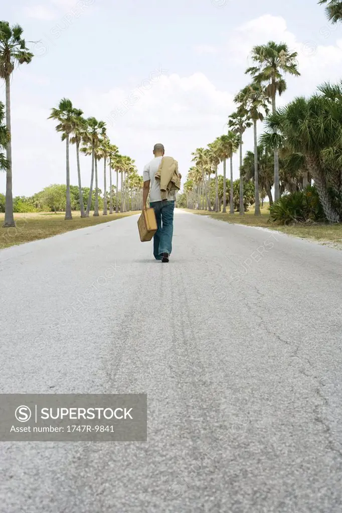 Man walking in center of road, carrying suitcase and jacket, rear view