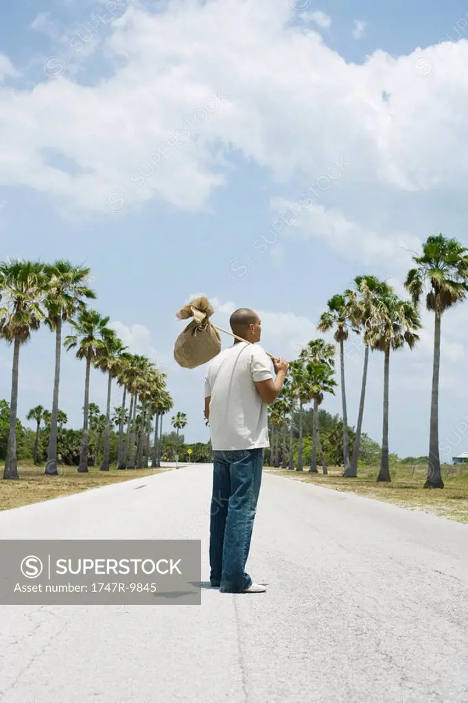 Man standing in middle of road, carrying burlap bundle on shoulder, rear view