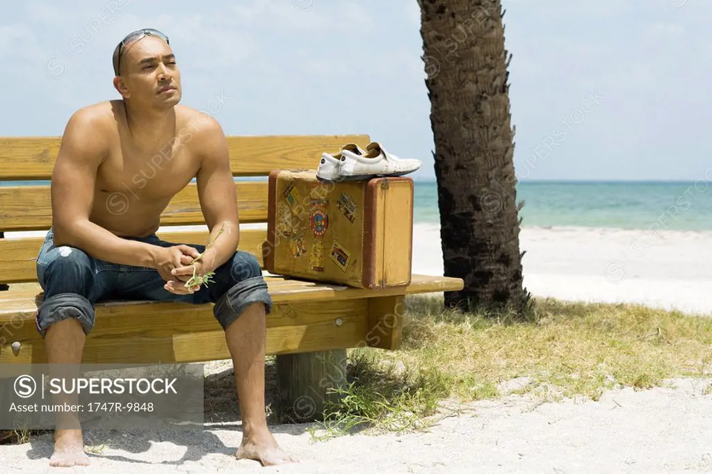 Man sitting on bench at the beach, suitcase beside him, looking away