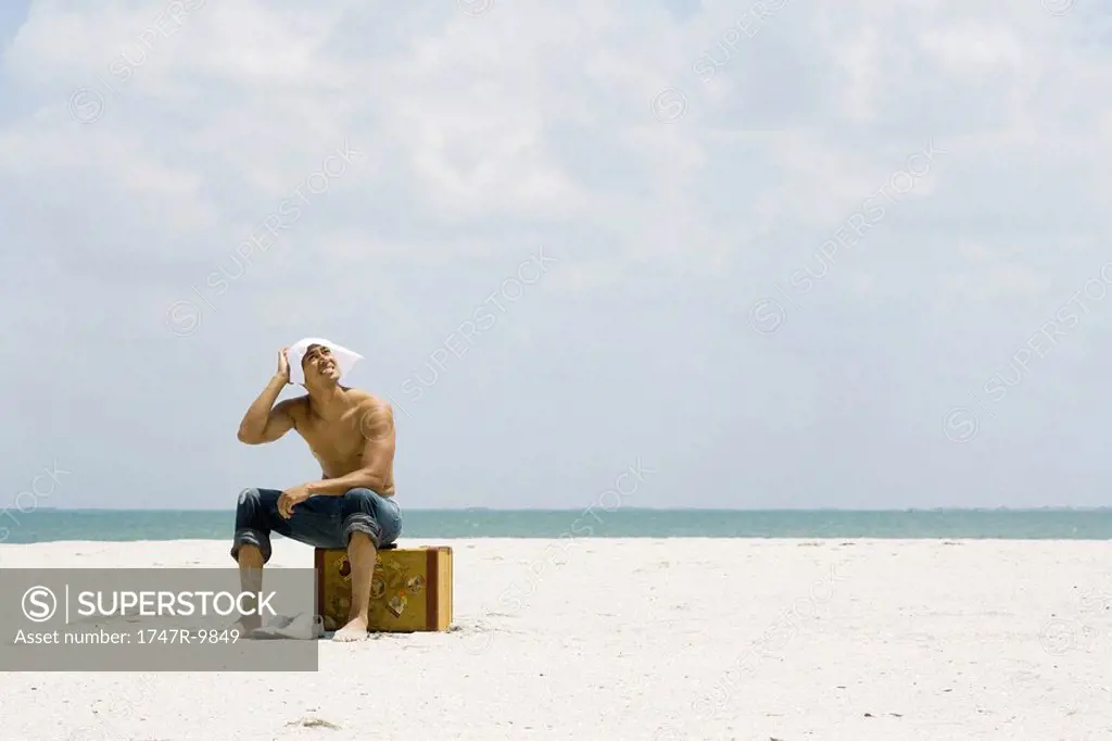Man sitting on suitcase at the beach, covering head with handkerchief, looking up at sun