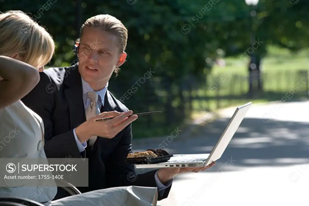 Businessman sitting on a park bench holding a laptop with a businesswoman sitting beside him