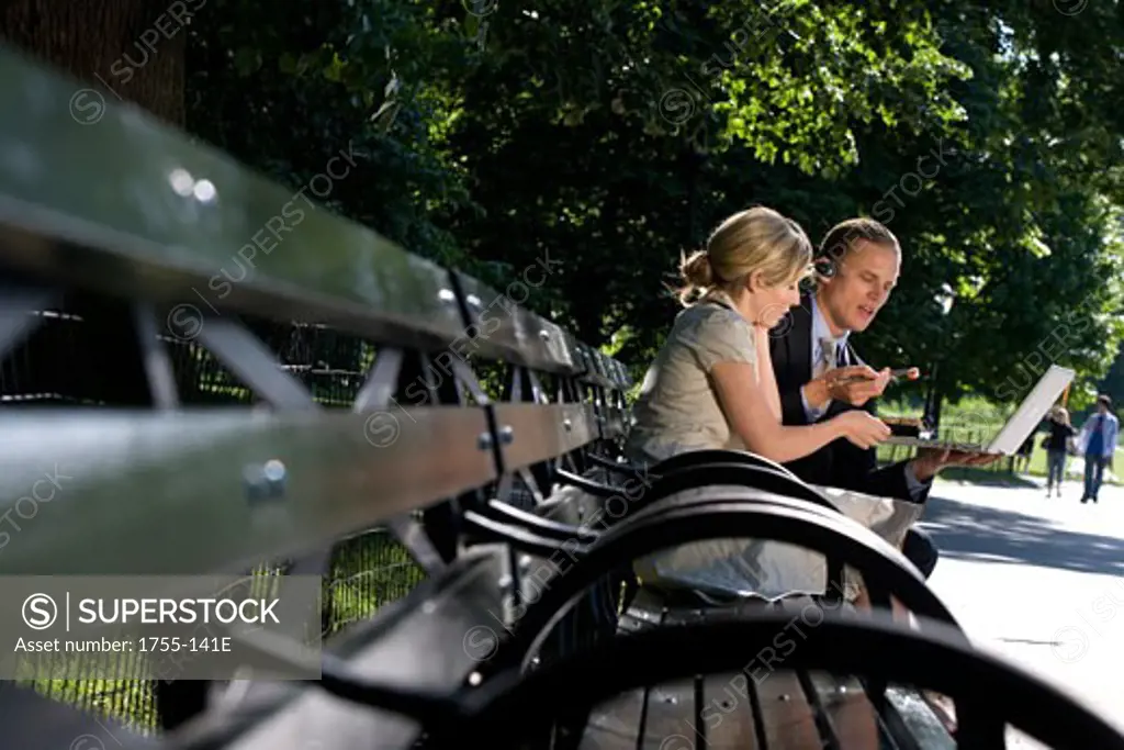 Businessman and businesswoman sitting on a park bench and looking at a laptop