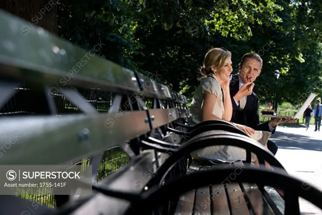 Businessman holding a laptop with a businesswoman sitting on a park bench and smiling