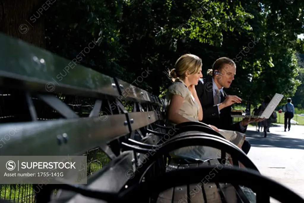Businessman and businesswoman sitting on a park bench and looking at a laptop