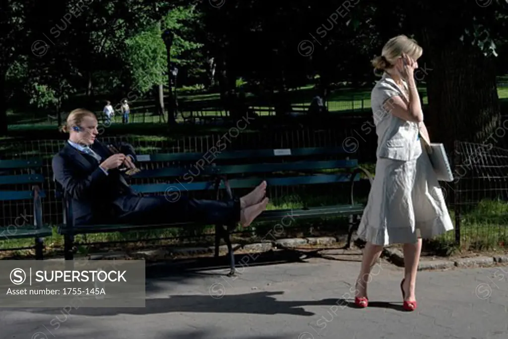 Businesswoman holding a laptop with a businessman reclining on a park bench behind her