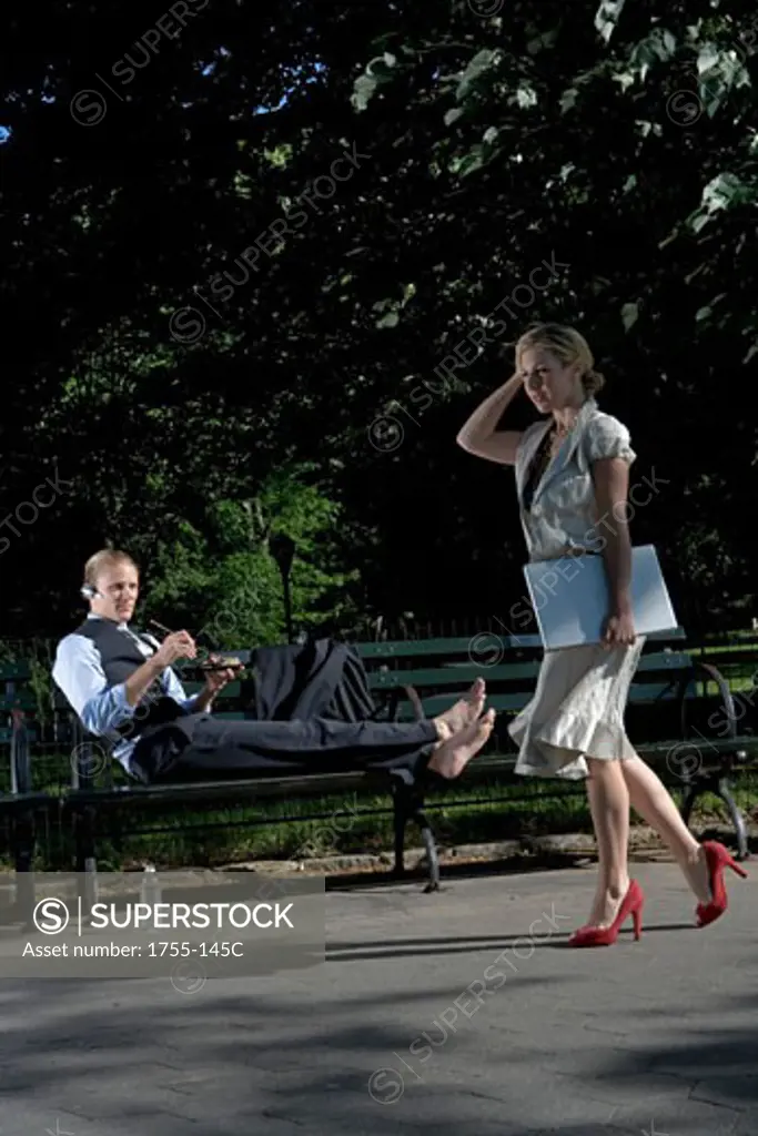 Businesswoman walking with a businessman reclining on a park bench