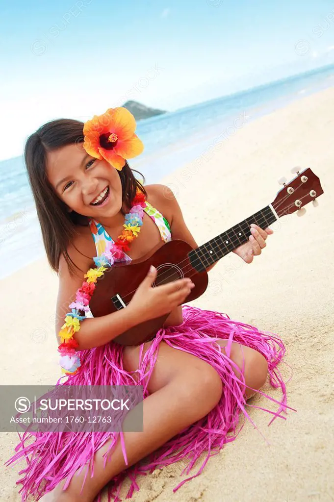 Hawaii, Oahu, Young girl smiling and playing ukulele on the beach in a hula skirt.