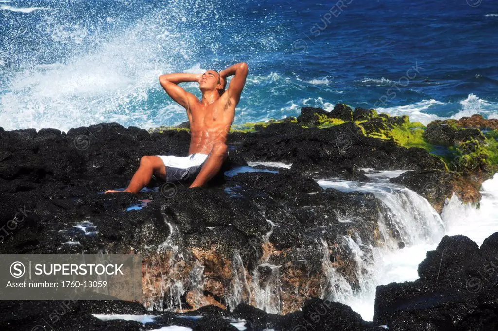 Hawaii, Oahu, Local young man enjoying sea mist on a sunny day on the rocks on shore.