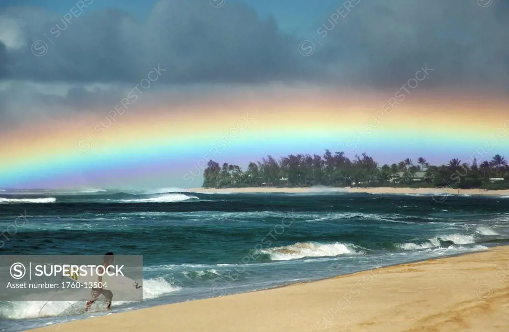 Hawaii, Oahu, North Shore, Surfer exiting water admires a beautiful, bright rainbow. Editorial Use Only.