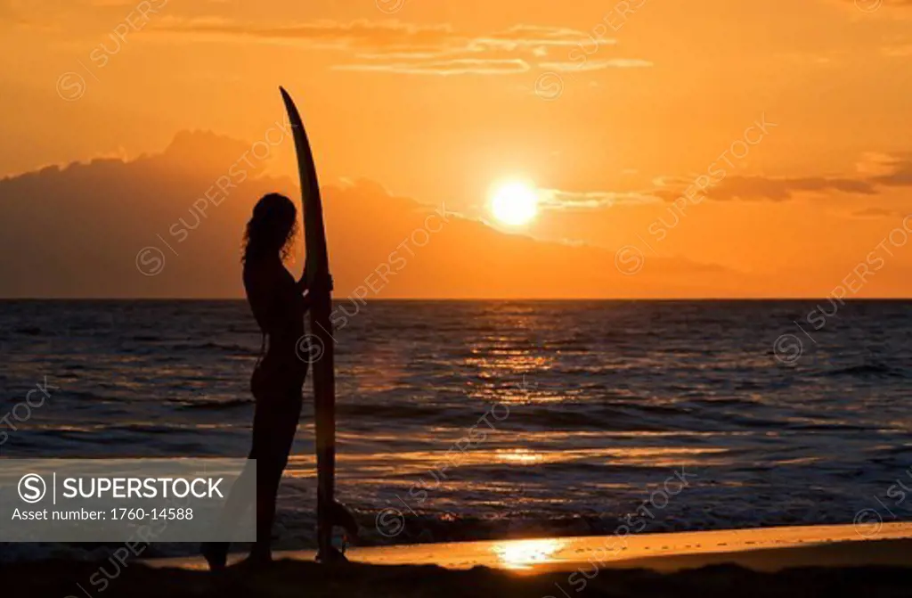 Hawaii, Female surfer on beach silhouetted against orange sunset over ocean.