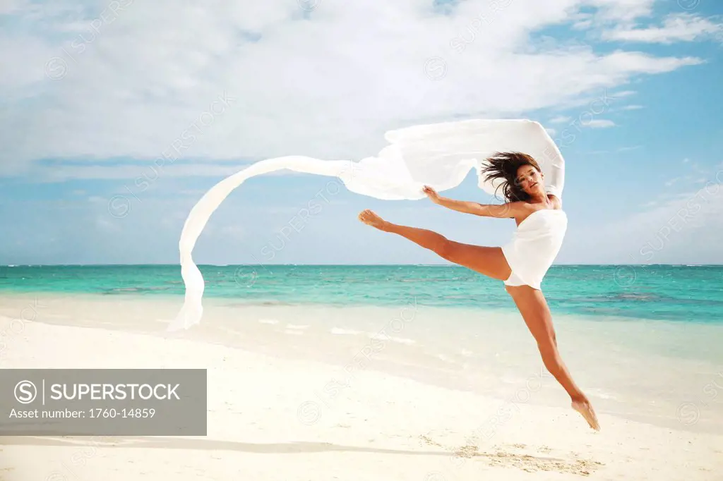 Hawaii, Oahu, Lanikai Beach, Beautiful female ballet dancer leaping into air on beach with white flowing fabric.