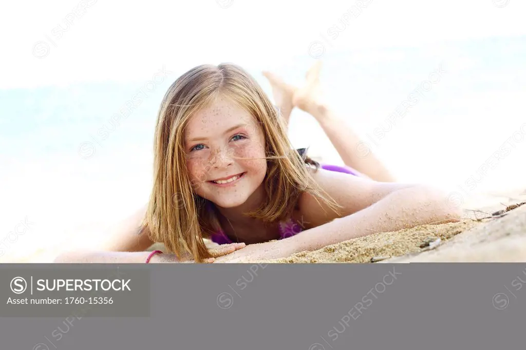 Young girl with red hair and freckles lays in the sand on the beach.