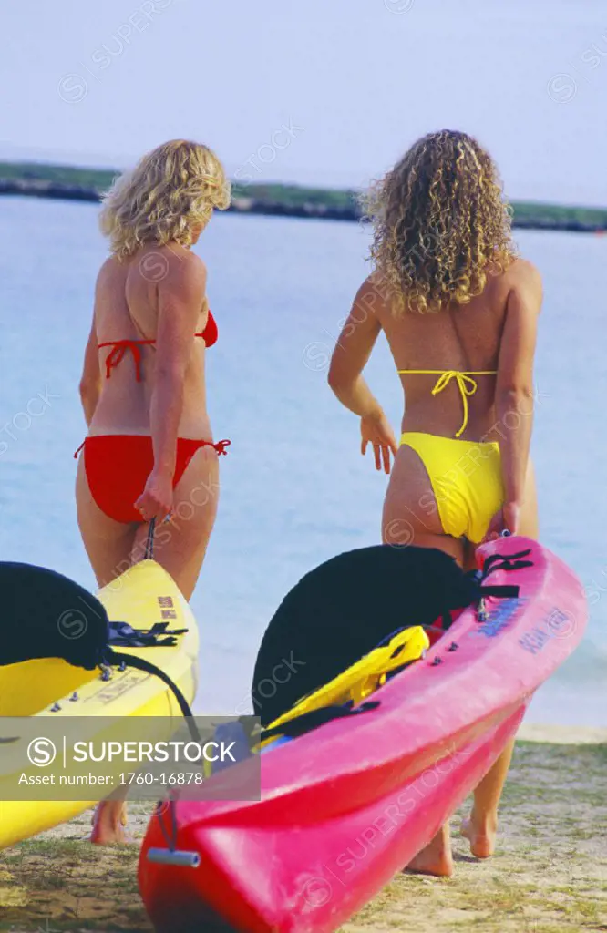 Two blonde women pulling kayaks into ocean, walking on sand.