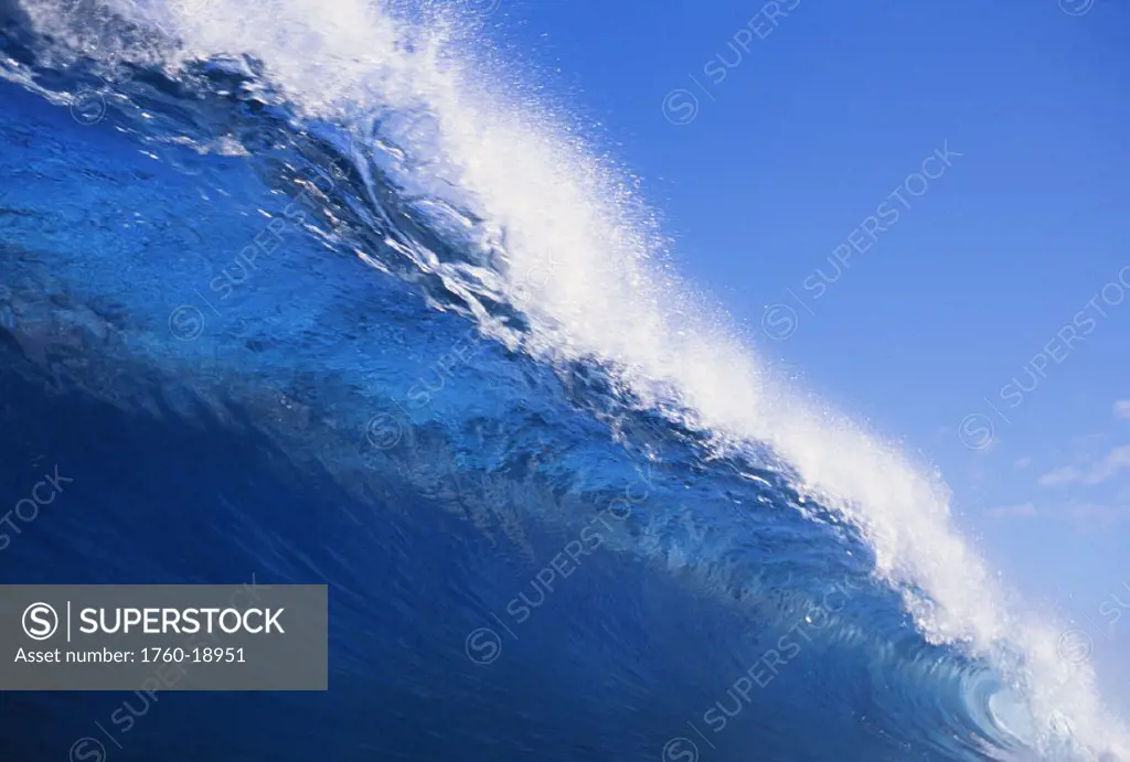 Closeup of translucent wave about to curl with spray, blue skies.