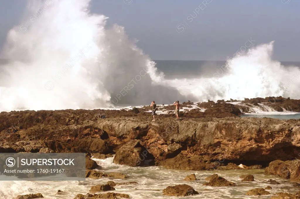 Hawaii, Oahu, North Shore, Sharks Cove, Two men stand on rocks photographing massive, crashing waves