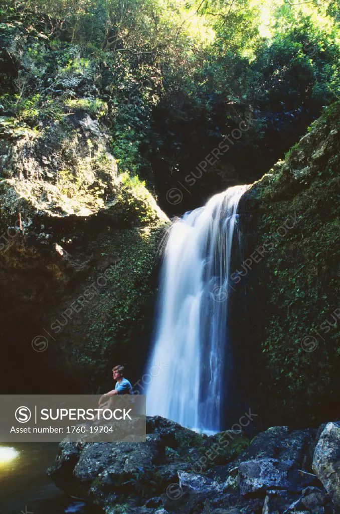 Hawaii, Maui, hiker rests on rock next to waterfall in Mokumokuele Valley