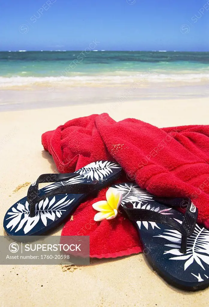 Pair of flip flops with towel and plumeria on white sand beach, blue ocean and sky in background